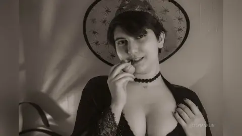 Moody black and white portrait of a woman in a dark lace outfit wearing a wide-brimmed hat indoors.