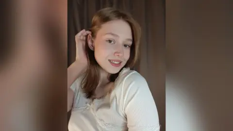 EvelinaMoorina smiles at the camera while running her hand through her light brown hair in a white top.