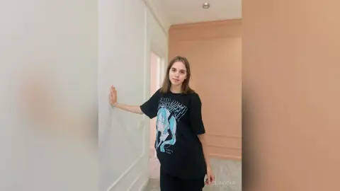 Young woman in a black graphic t-shirt standing in a bright, minimalist white hallway with a calm mood.