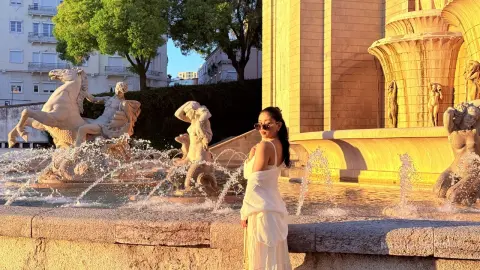 Woman in white summer outfit smiling by a classical stone fountain in golden hour sunlight.