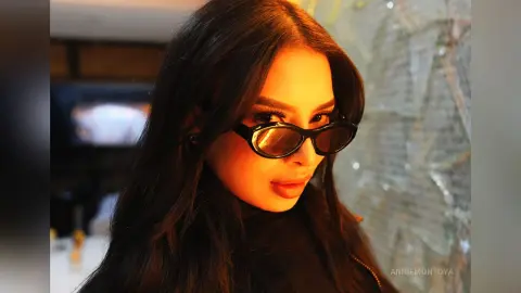Stylish woman with long dark hair wearing trendy black sunglasses in a warm, moody indoor setting.