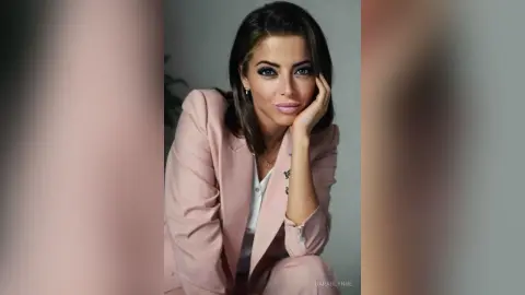 Elegant woman in a blush pink blazer posing with a thoughtful expression against a neutral studio background.
