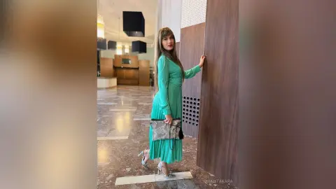 QuinnTakara poses in a long teal dress and silver heels while standing next to a wooden wall in a modern lobby.