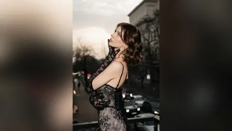 Elegant woman in a black lace dress and gloves posing on a city balcony during a moody sunset.