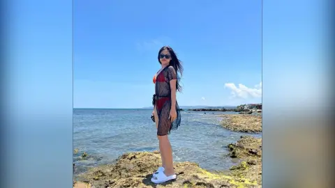 Stylish woman in a black sheer cover-up and sunglasses standing on rocky shores by the blue ocean under a sunny sky.