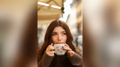 Young woman in a cozy brown sweater sipping coffee from a floral mug at an outdoor cafe on a sunny day.