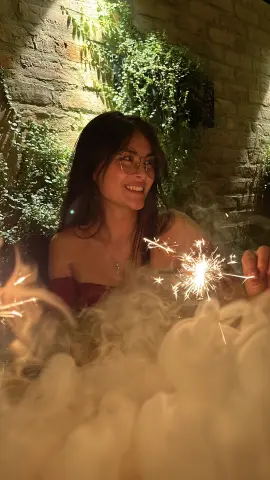 Smiling woman in glasses holding a sparkling firework against a rustic brick wall background at night.