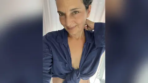 MichaelaBelle wears an unbuttoned blue linen shirt, looking toward the camera with her hand behind her neck.
