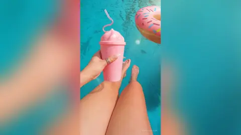 Person holding a pink milkshake by a swimming pool with a floating pink donut on a sunny summer day.