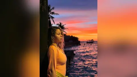 Young woman in an orange top gazing at a vibrant sunset over the ocean during a peaceful tropical evening.