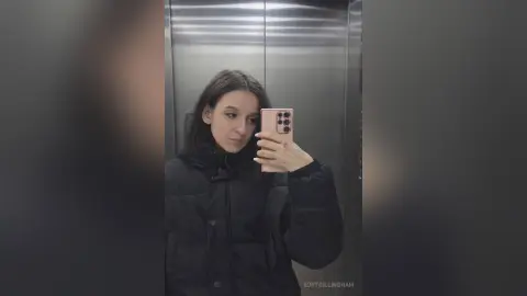 Young woman in a black puffer jacket taking a moody mirror selfie inside a modern elevator.