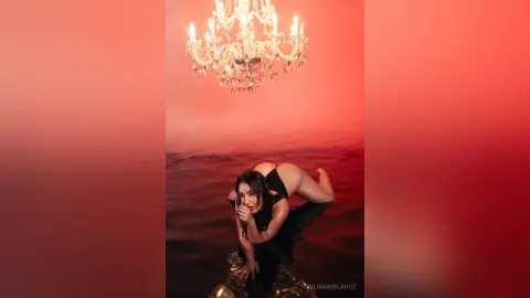 JulianaDeLaHoz poses naked in a black outfit, arching her back and showing her bare buttocks while leaning over a gold chair under a lit chandelier.