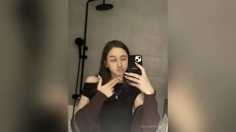 Young woman in a black off-the-shoulder top taking a playful mirror selfie in a modern bathroom.