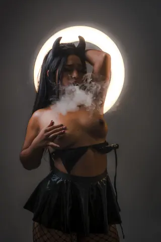 Mystical woman in black devil costume with horns posing amidst ethereal white smoke in a moody studio setting.