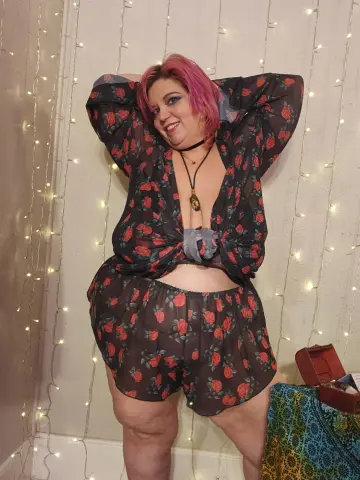 Smiling woman in floral black loungewear posing against fairy lights in a cozy indoor setting.