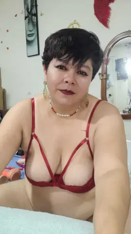 Woman with short dark hair wearing red lingerie and pearl necklace in a bright indoor setting.