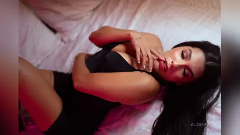 Young woman in a black dress lying on white bedding with a moody, romantic atmosphere.