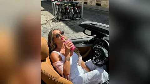 Blonde woman in white tank top sipping a pink drink while relaxing in a convertible car on a sunny day.