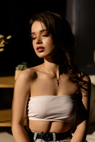 Young woman in a white tube top and silver cross necklace posing in warm, moody sunlight.