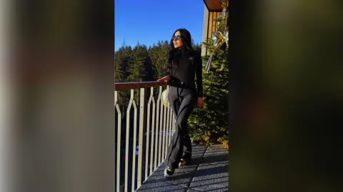 Stylish woman in black outfit and sunglasses standing on a sunny balcony overlooking a pine forest.