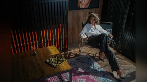 Stylish woman in white blouse and black trousers sitting in a sunlit room with a wooden table and chessboard.