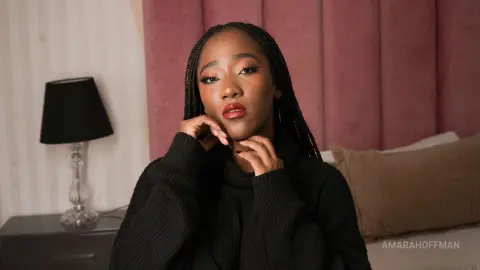 AmaraHoffman with long braids wearing a black sweater, posing in a bedroom setting.