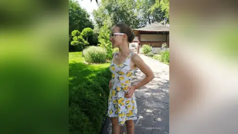 Smiling woman in a yellow floral sundress standing on a stone path in a sunny garden setting.