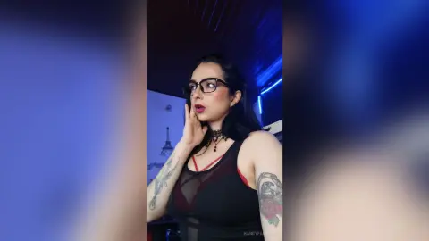 Tattooed woman in a black tank top posing under moody blue lighting in a modern room.