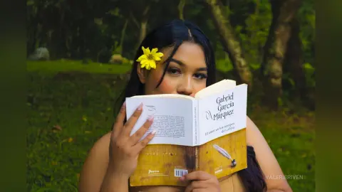 Young woman with a yellow flower in her hair reading a Gabriel García Márquez book in a lush green forest.