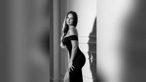 Elegant woman in a black off-the-shoulder dress posing in a moody, minimalist indoor setting.