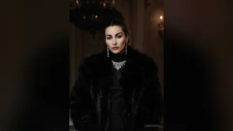 Elegant woman in a black faux fur coat and diamond necklace posing in a moody, luxury indoor setting.