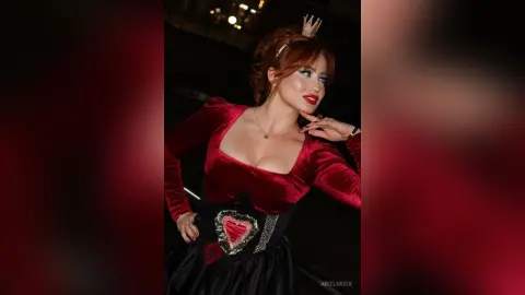 Elegant woman in a red velvet gown and black corset posing with a regal expression in dim lighting.