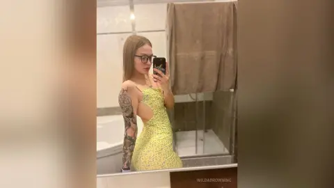Young woman with arm tattoos wearing a yellow floral cutout dress taking a mirror selfie in a modern bathroom.