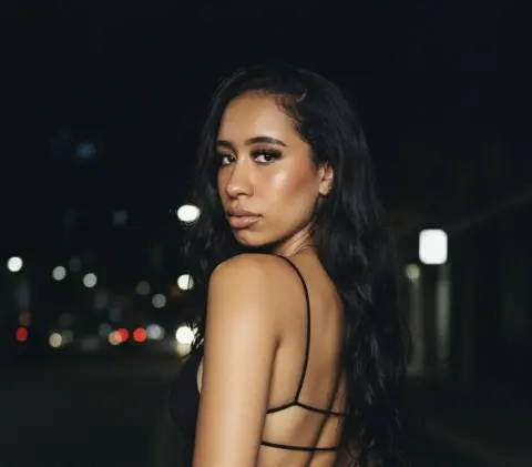 Young woman in a black strap top posing with a moody expression against a dark city night background.