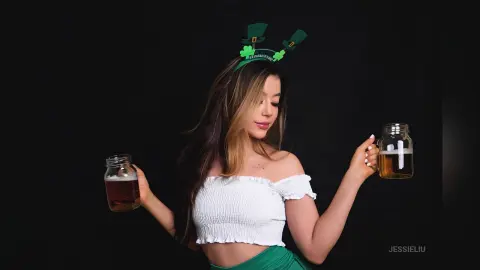 JessieLiu holds a glass mug of beer in each hand while wearing a white off-the-shoulder top and green headband against a black background.