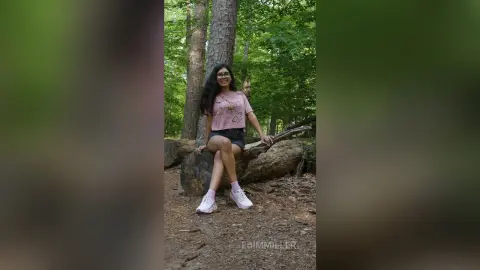 Smiling woman in a pink t-shirt and denim shorts sitting on a rock in a lush green forest.