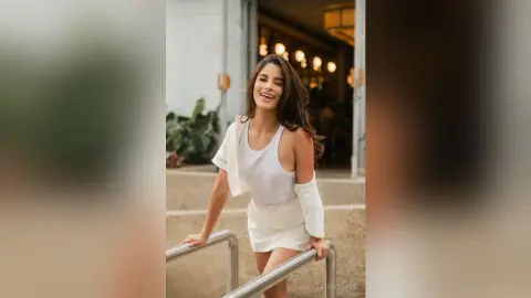 TefyRusso smiles while wearing a white tank top and white skirt, posing outdoors next to a metal handrail.