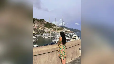 Woman in floral summer dress looking at boats in a scenic Mediterranean coastal harbor at sunset.