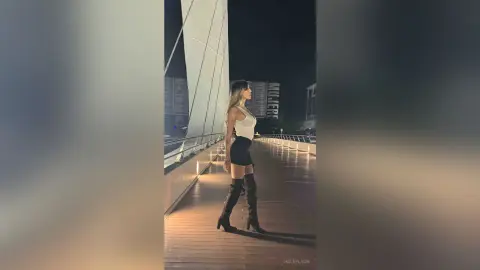 Stylish woman in a white tank top and black mini skirt posing on a wooden pier at night.