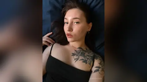Young woman with tattoos in a black tank top lying on a dark blue bed with a moody, relaxed expression.