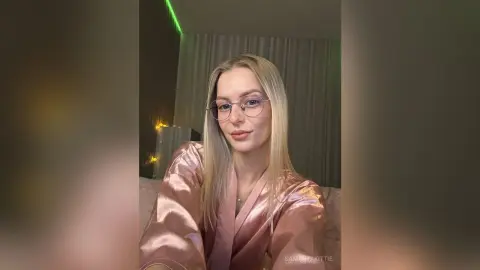 Blonde woman in a pink satin pajama set smiling in a cozy, dimly lit bedroom setting.