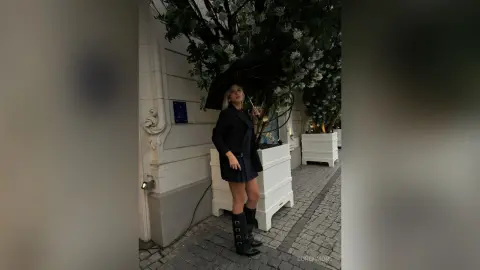 Stylish woman in a black blazer and mini skirt holding a black umbrella on a city street near white planters.