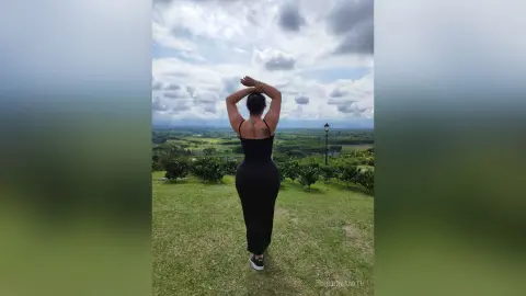 Woman in a black maxi dress standing in a green field under a cloudy sky, enjoying a peaceful landscape view.