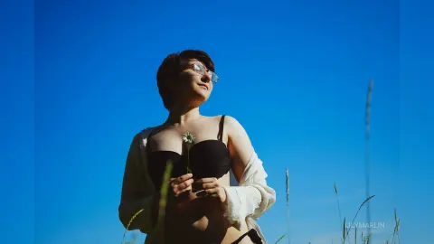 Young woman in a white cardigan and black bikini top standing under a clear blue sky in a sunny meadow.