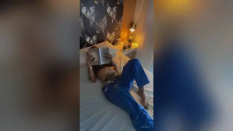 Relaxed woman in blue loungewear reading a book on a white bed in a cozy, dimly lit bedroom.