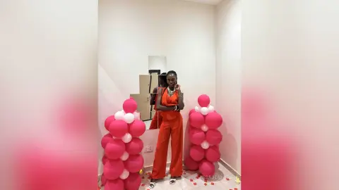 Stylish Black woman in a vibrant red jumpsuit posing next to pink balloon decor in a modern white room.