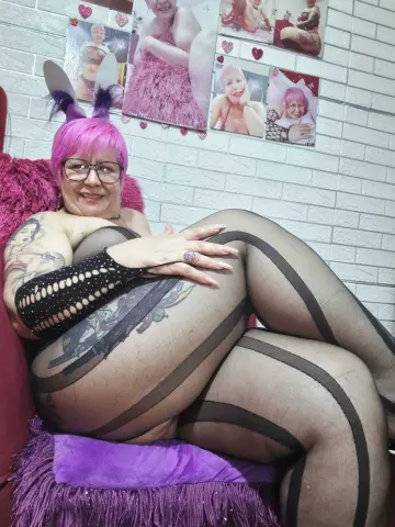 Smiling woman with pink hair and tattoos wearing black patterned tights on a purple faux fur chair.