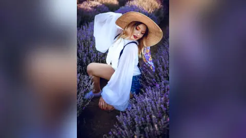 AmelieSkylar wears a white long-sleeve blouse and denim shorts while sitting in a field of purple lavender wearing a wide-brimmed straw hat.