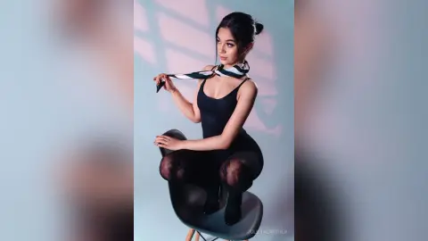 Elegant woman in a black bodysuit and patterned scarf sitting on a chair in soft cinematic lighting.