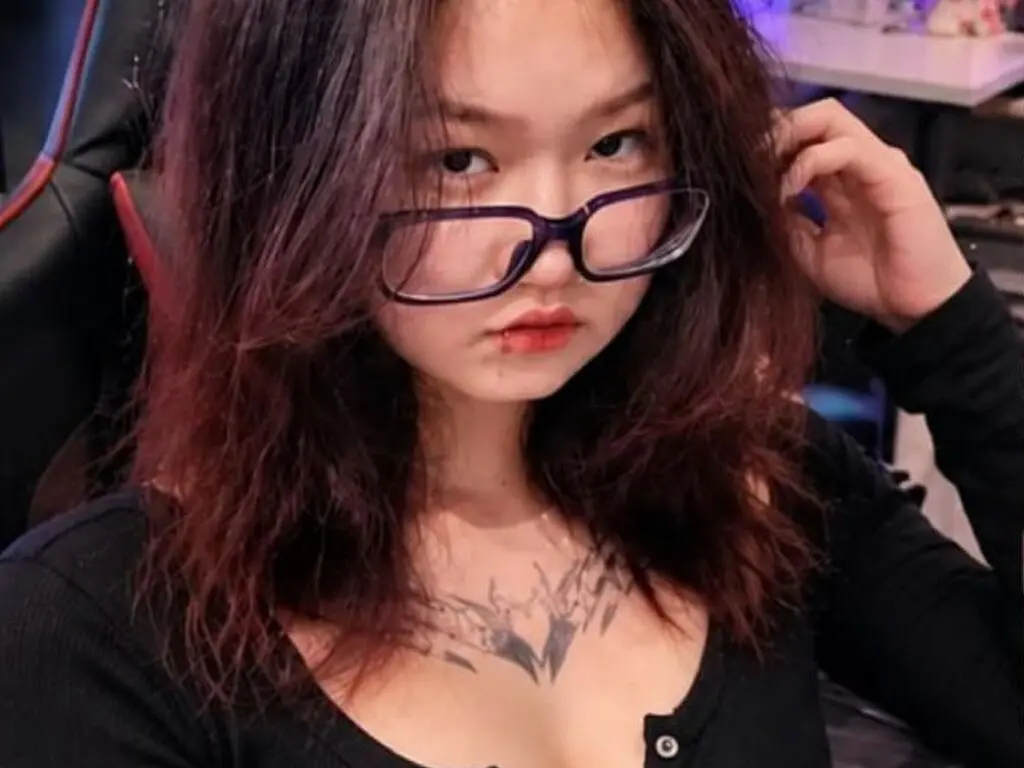 TishaOyervides wearing glasses and a black top, showing her long reddish-brown hair and chest tattoo while sitting in a chair.